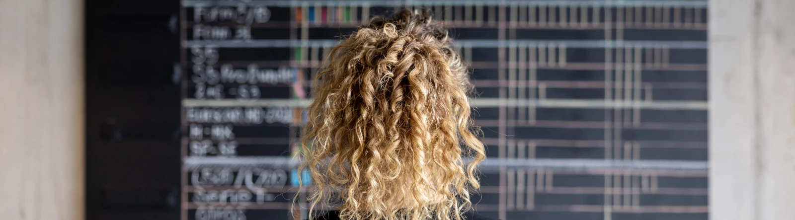 A woman with curly blonde hair looking at a calendar on a blackboard