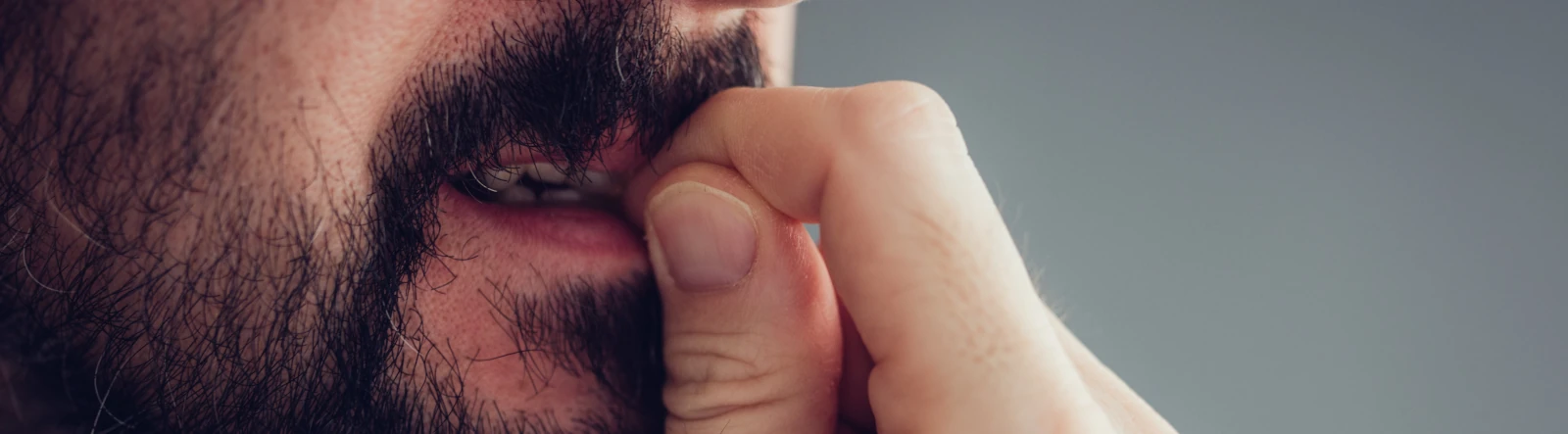 The lower half of the face of a bearded man biring his fingernails