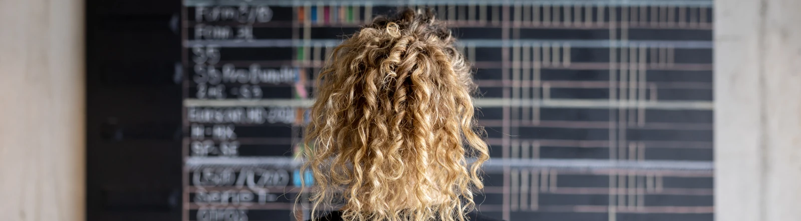 The back of the head of a woman with curly blonde hair looking at a calendar on a blackboard