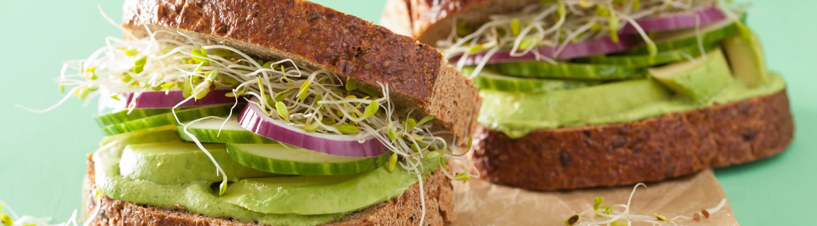 Two sandwiches filled with avocado, cucumber, red onion, and cress